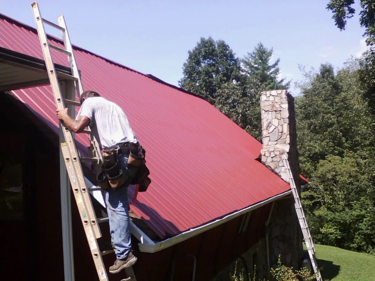 Roofer on a ladder beside a red metal roof and stone chimney — Soffit & Fascia Repair by Roofing Brothers in Providence